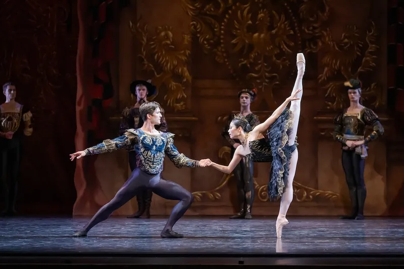 a man and a woman dancing ballet; woman is japanese origin, in a black dress with her weight on one foot and her other leg balanced above in a split