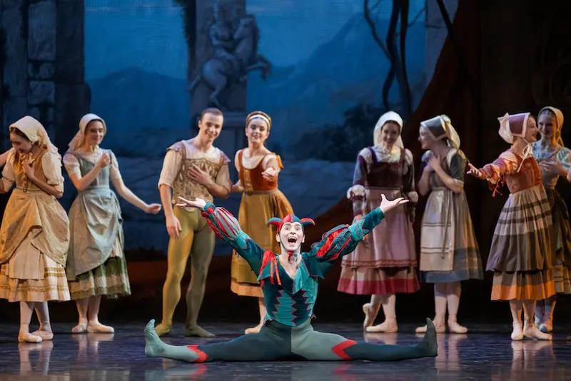 a man in a green and red jester outfit grins while straddling the stage while pairs of dancers in peasant costumes look on
