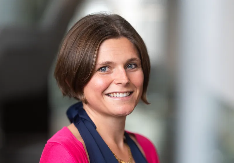 a white woman with light brown hair, wearing a blye and pink top and a smile, soft focus