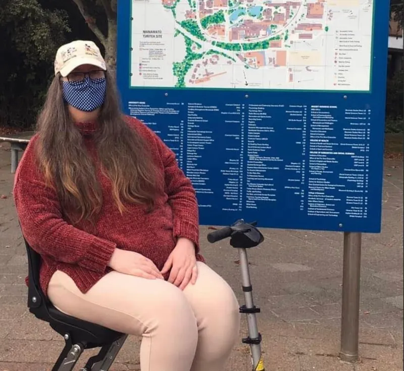 a woman in a motorised wheelchair with long brown hair and a facemask, wearing a red sweater and in front of a blue map
