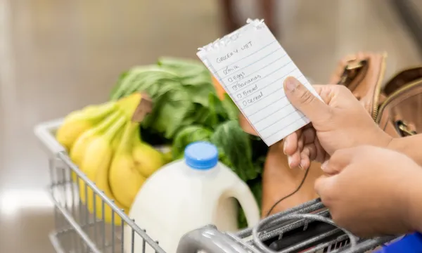 a hand holding a shopping list above a shopping trolley that contains milk bananas and spinach