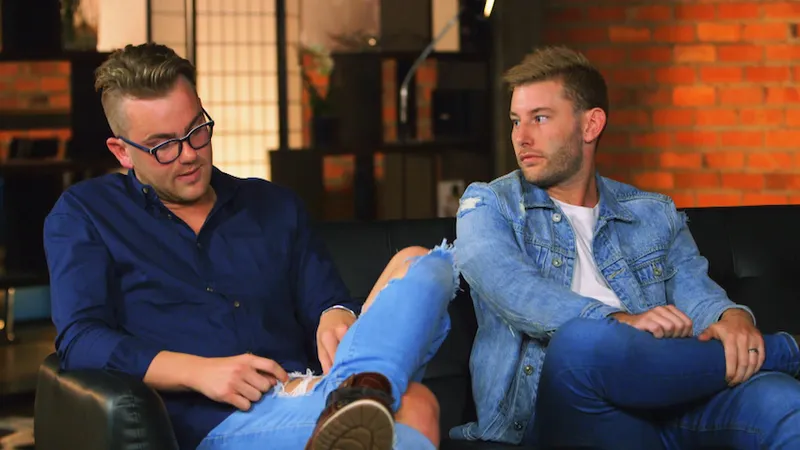 Two blonde men sit on a leather sofa looking annoyed at each other