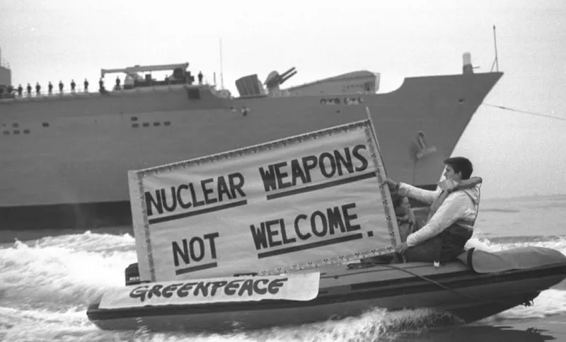Person holding banner saing 'Nuclear weapons not welcome' on a little boat in front of a large boat