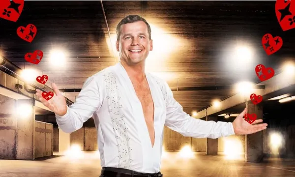 bright lights in the background, a man with lots of fake tan holds his arms wide while wearing a white shirt with a deep v cut to his bellybutton, and a slightly unsettling look in his eyes