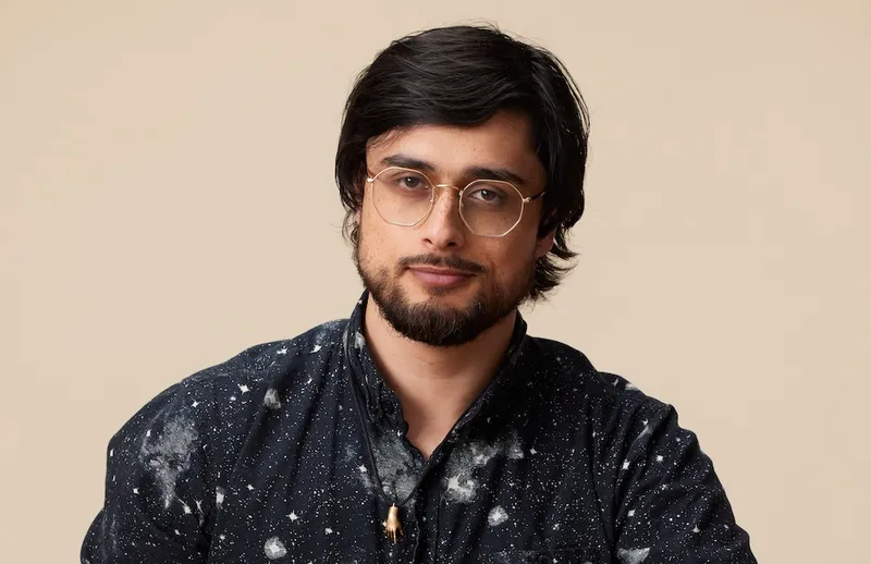 a man with light brown skin, , a slightly scruffy black beard, glossy hair around his airs, clear acetate glasses, and a textured navy shirt