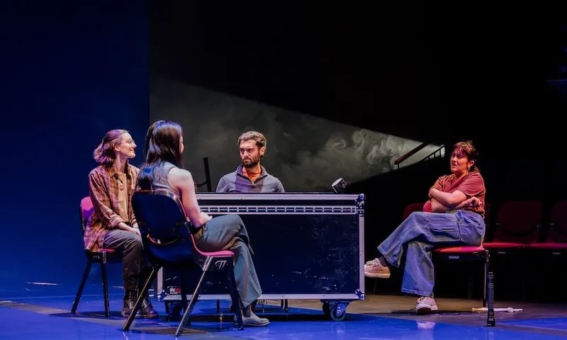 a blue-lit stage with five actors sitting around a table, some smoke drifting through the spot lights so they have more dimension. It looks a little gloomy, and certainly intriguing