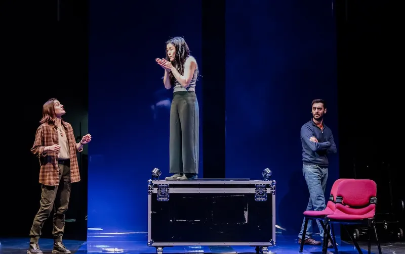 Dawn Cheong, a short Asian woman wearing flowy grey pants and a sleeveless top stands on a black box while two characters on each side of the blue-lit stage look on