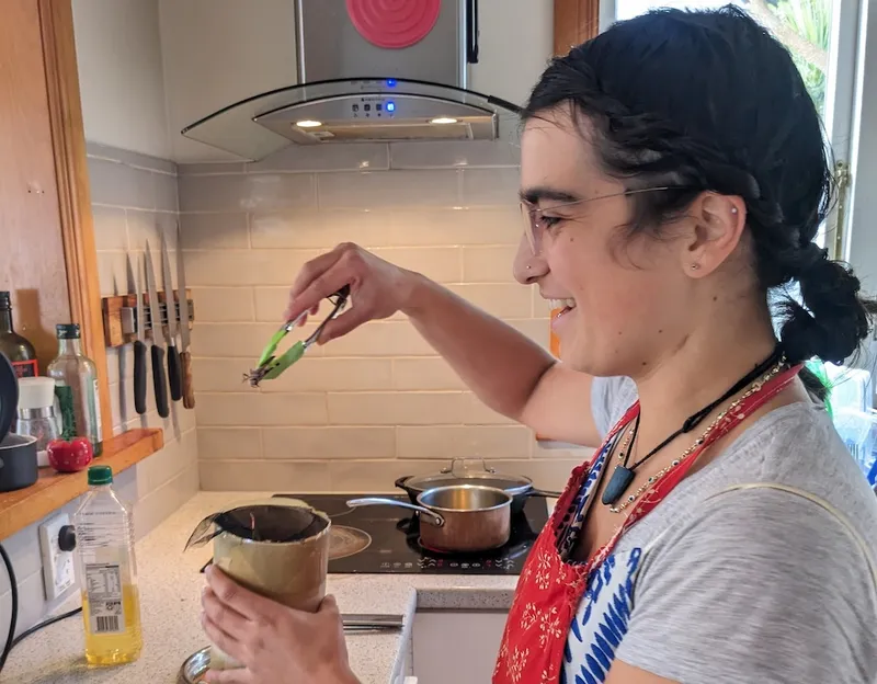Shanti, a woman with light brown skin, glasses, and a grey t-shirt, grins nervously as she pulls a frozen locust out of a cardboard tube with a pair of green tongs. The stove with a hot, oily pan waiting in the background is a sign of the locust's eventual fate