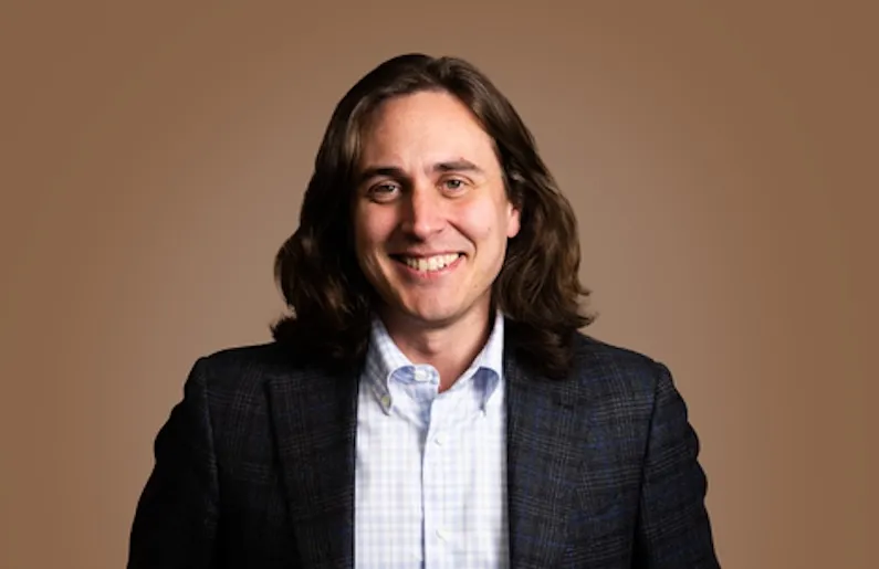 a brwon background with a white man with glossy long dark brown hair wearing a suit jacket and shirt and smiling at the camera