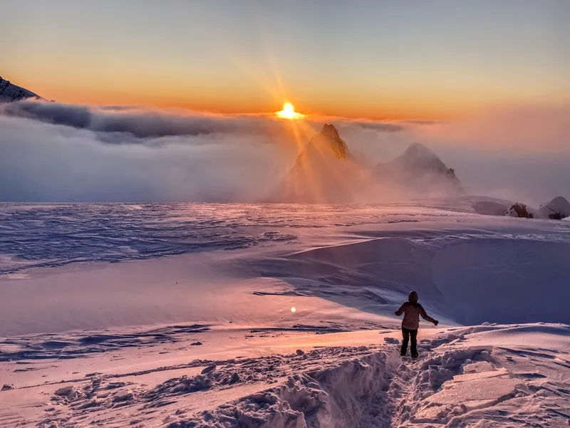 the sun setting over the mountains with a big field of snow and a figure appropriately decked out in warm and waterproof clothing walking through some curned up snow