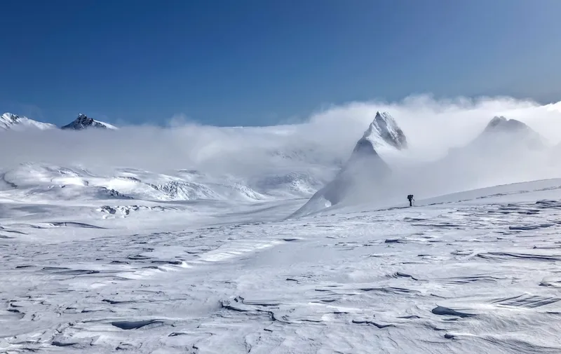 a snowy day in the mountains that makes you wish you had crampons on and were in the big wide world not looking at a screen; the sky is clear and blue, little cloaks of cloud are draped around the maunga, and a figure walks through a snow field in the sunshine