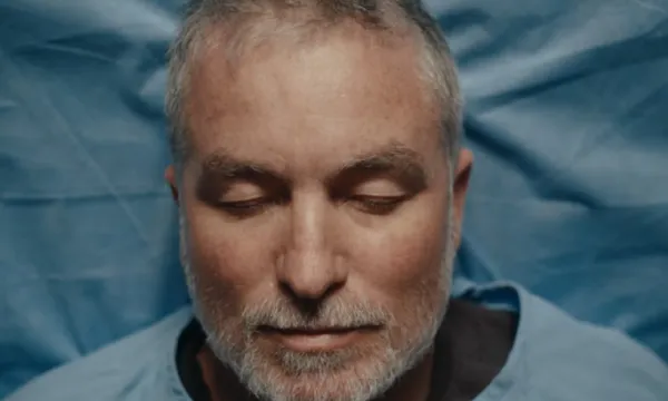 A close-up of Dai Henwood lying on his back with his eyes closed. He is resting on a hospital bed or surgical table, wearing a surgical gown. It is a still image from Dai henwood's TV show, Live and Let Dai on Three.