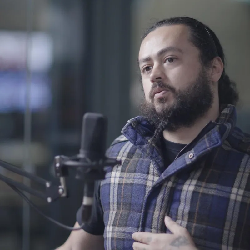 A man with a beard and long hair speaking into a microphone, wearing a blue flannel shirt.