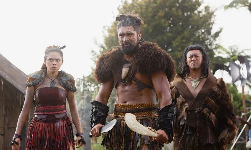 Three people in traditional warrior attire, with fierce expressions, stand outdoors. The central figure holds a weapon, while all wear furs and leather, surrounded by trees and huts in the background.