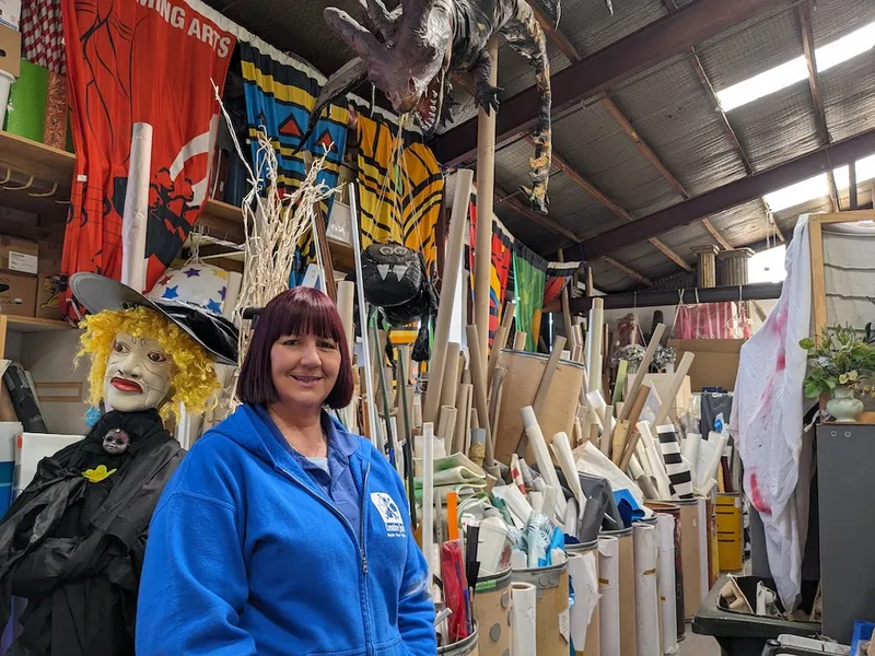 Christine Gayton, a middle aged woman with dyed red hair and a bright blue jacket smiles at the camera with a delightful chaos of cardboard tubes, a mannequin witch, and a giant dinosaur behind her