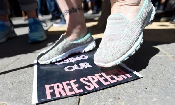 A person steps on a sign on the ground that reads "USING OUR FREE SPEECH" during a protest or demonstration. Other people's feet are visible in the background.