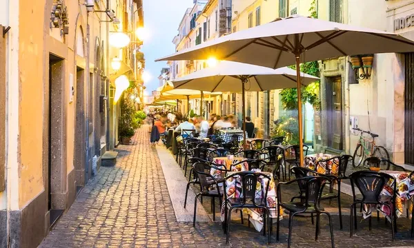 A cozy outdoor café in a European street with cobblestones and warm lights. Empty chairs and tables with colorful cushions line the walkway. People are dining under large umbrellas against a backdrop of charming, rustic buildings.