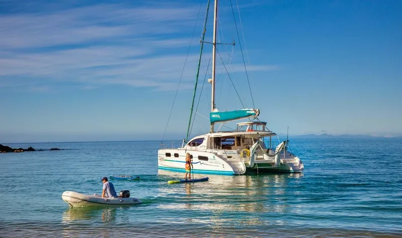 a sunny day with turquoise water and a sail boat, with a woman paddleboarding in the centre of the picture, and a man in a rubber dinghy with an outboard motor puttering towards shore at the edge of the frame