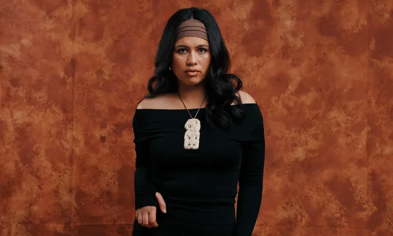 A woman with long, dark hair wears a black off-the-shoulder top and a headband. She has a large pendant necklace. The background is a textured brown backdrop.