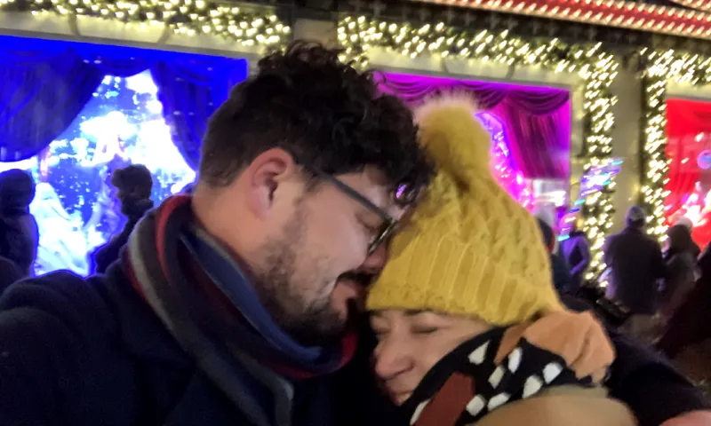 A couple embraces warmly in front of a festive store window.