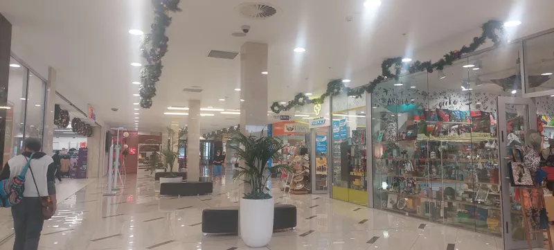 A shopping mall corridor decorated with holiday garlands. Features various stores, including a toy shop with colourful displays and a travel agency. Shoppers walk through the area, and potted plants are placed along the tiled floor.