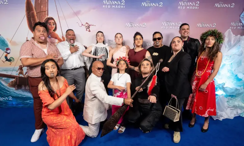 A group of people dressed in traditional Maori attire pose playfully on a blue carpet for a "Moana 2 Reo Māori" event. The backdrop features animated scenes from the movie. Some individuals make expressive faces and gestures.