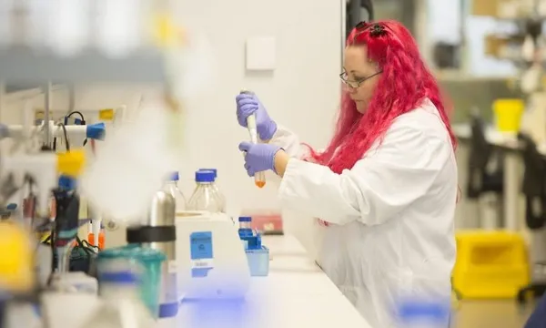 a pink haired white woman wearing a lab coat and gloves, putting something in a bottle with a pippette