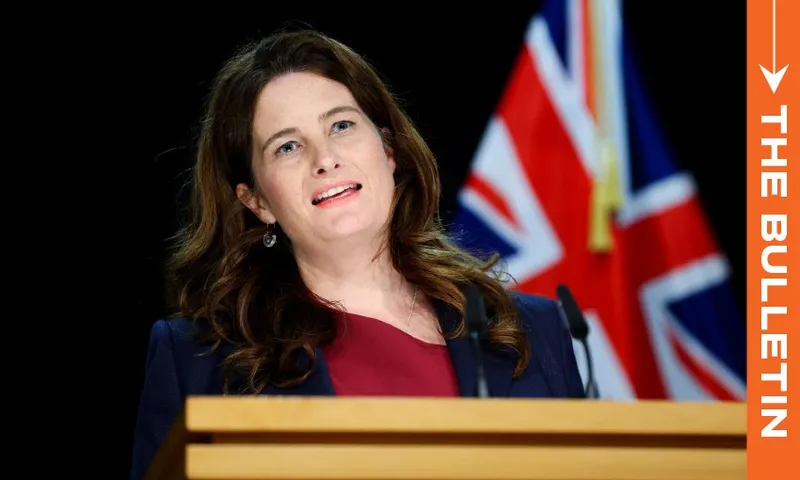 A woman is speaking at a podium with a microphone, wearing a dark blazer and red top. A flag with red, white, and blue colors is visible in the background. The image has a vertical orange banner on the right with the text "The Bulletin.