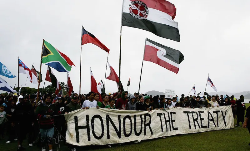 A large group of people hold flags and a banner reading "HONOUR THE TREATY" during a protest or demonstration outdoors on a cloudy day.