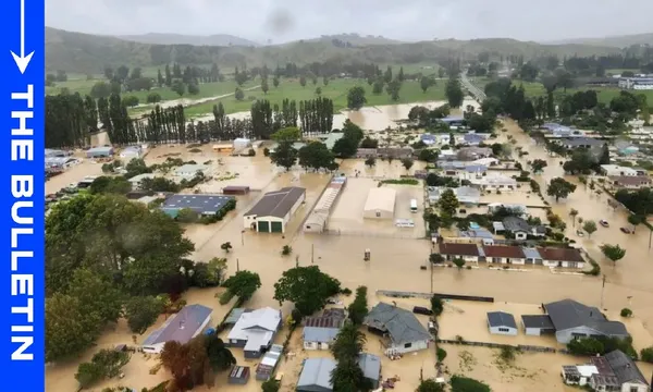 Aerial view of a flooded town with muddy water covering streets, homes, and buildings; trees and green hills are visible in the background under a cloudy sky. “THE BULLETIN” is written vertically on a blue bar at the left edge.