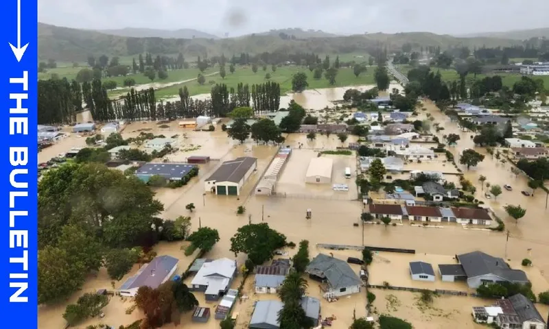 Aerial view of a flooded town with muddy water covering streets, homes, and buildings; trees and green hills are visible in the background under a cloudy sky. “THE BULLETIN” is written vertically on a blue bar at the left edge.