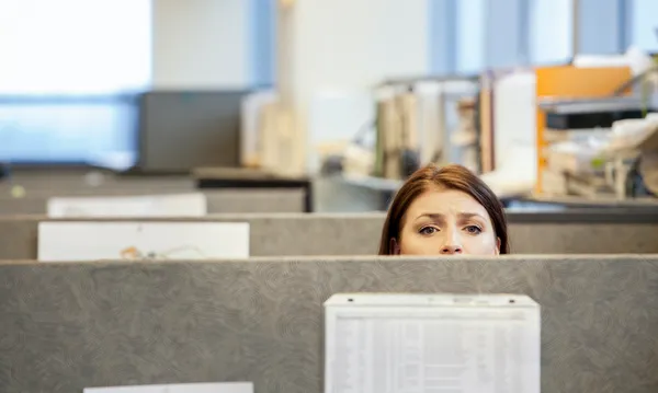 A young Caucasian woman looking concerned over the top of her cubicle in a corporate office.