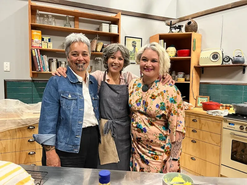 Tania Gilchrist, Kim Garrett and Whiti Hereaka huddle together and smile for a photograph in the onstage kitchen set.