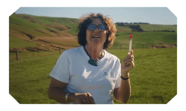 Dr Bev Lawton holds up a test tube in front of some rolling green hills of rural Otago