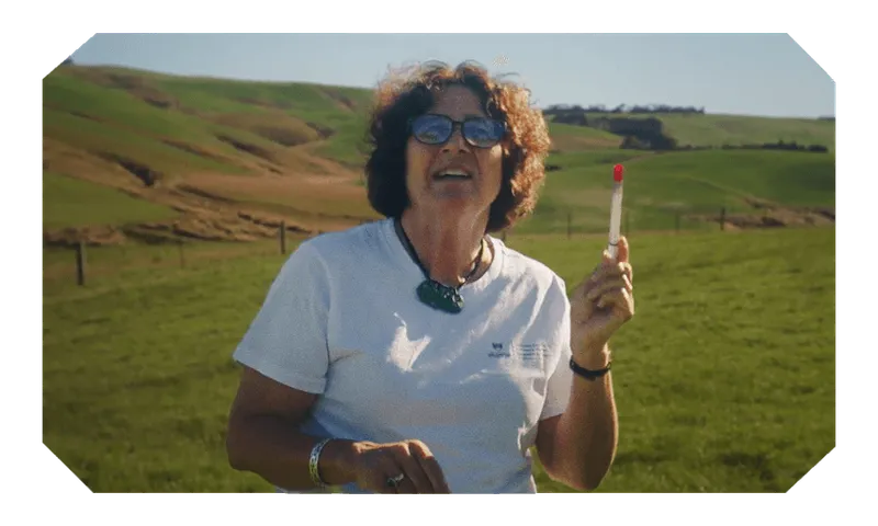 Dr Bev Lawton holds up a test tube in front of some rolling green hills of rural Otago