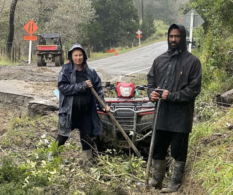 two Tokomaru residents dressed in wet weather gear holding shovels on the side of a road amid storm debris