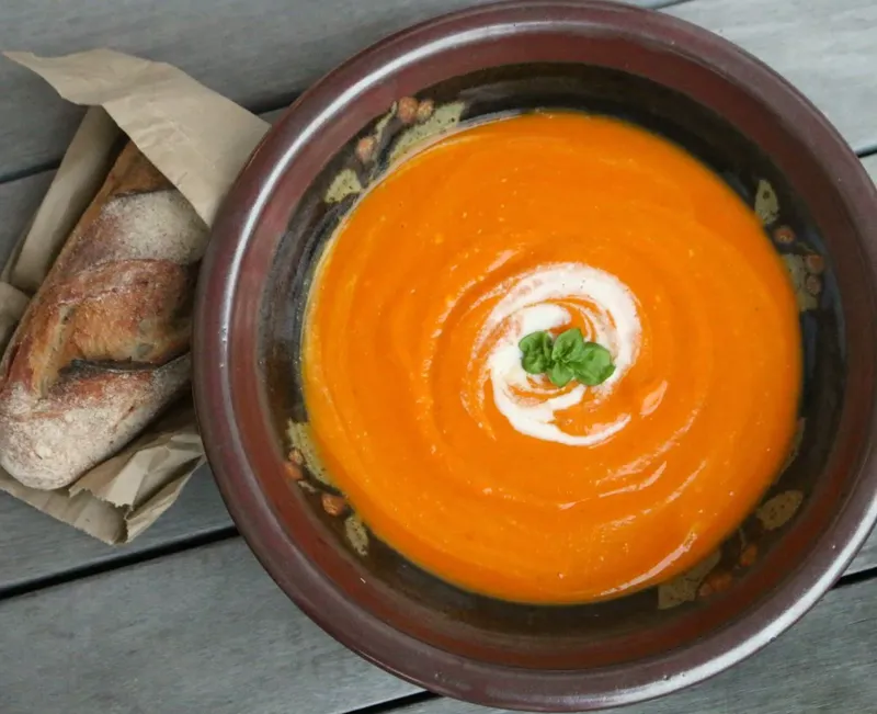 A bowl of smooth pumpkin soup topped with basil. On the side is a chunk of baguette.
