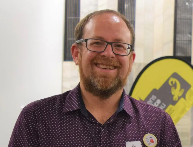 a midle aged white man with glasses and a purple shirt - a "I heart CAB" badge prominent on his chest