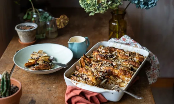 on a wooden table sits a baking dish filled with pear and chocolate croissant pudding. To the left is a serving of the pudding in a bowl, with a jug of cream next to it. There are flowers and plants and a red napkin on the table.