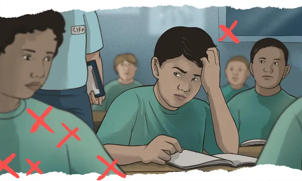 a boy is sitting at a school desk looking anguished, with his hand on his head. An adult wearing a CYFS badge approaches