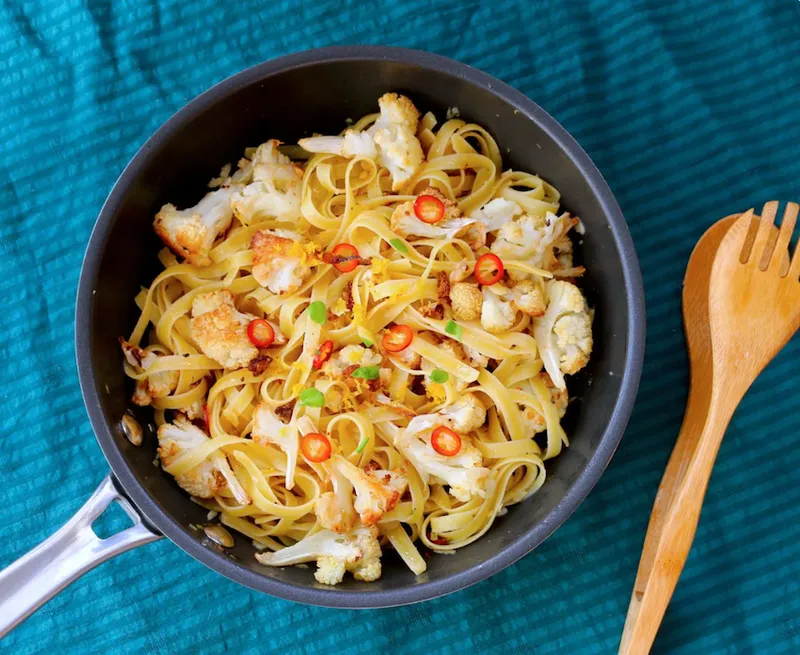 A bowl of roasted cauliflower pasta with flecks of fresh red chilli. The bowl is atop a blue tablecloth.