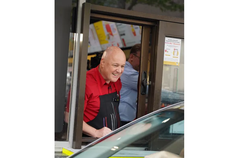 National party leader Christopher Luxon wears a red polo and a black apron as he leans out the window of a McDonald's drive through to talk to a customer..