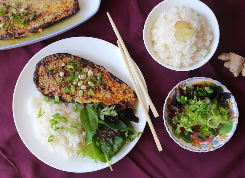 Birdseye view of a plate fillled with half a grilled eggplant, rice and a green salad. A bowl of rice and a bowl of salad along with chopsticks sit alongside on a purple tablecloth.