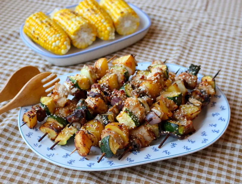 A plate piled with chicken and courgette skewers. Just behind is a plate with four grilled corn cobs. This is all served atop a brown and white gingham tablecloth.