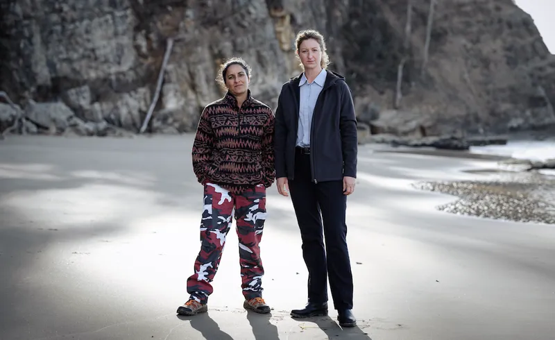 Two characters from the crime show Deadloch stand on a beach.