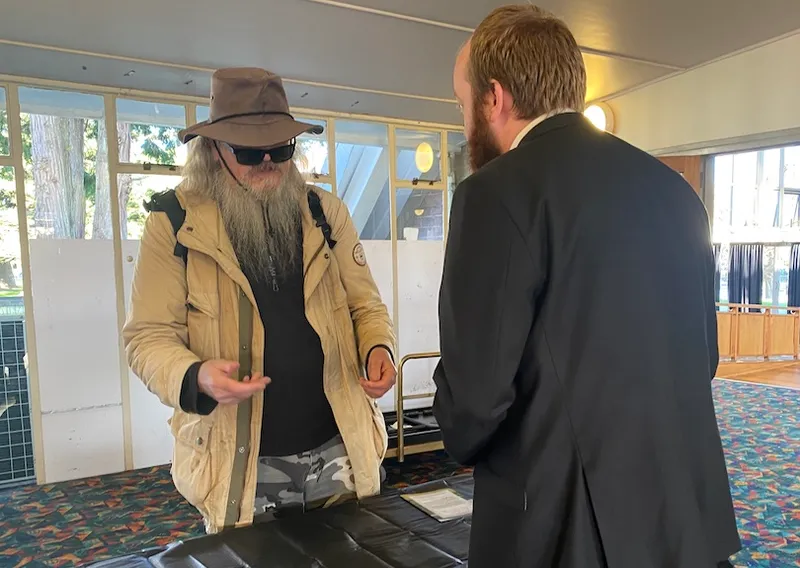 A man with a long grey beard, wearing a wide-brimmed hat secured beneath his chin, sunglasses a jacket, a backpack and camo trousers, talks to a man in a suit behind a desk