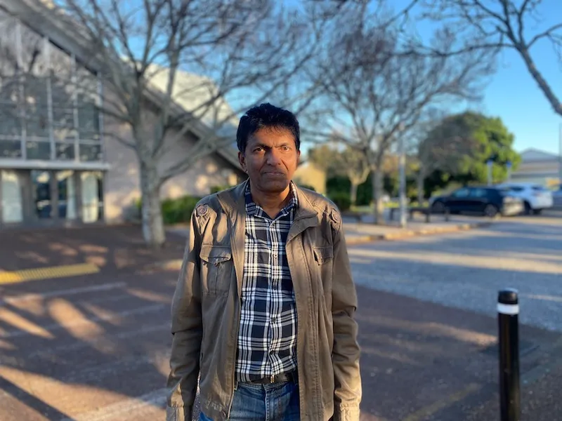 A Sri Lankan man in a checked shirt and brown jacket stands in front of a pyramid-shaped modernist building and a row of leafless trees