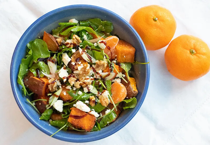a blue bowl filled with with greens, kūmara chunks, feta and mandarin segments. There are two mandarins sitting next to the bowl.