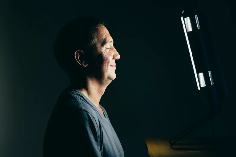 Smiling middle aged man sitting in front of a light therapy box, a full spectrum light box which mimics the sun, and treats people suffering from seasonal affective disorder.