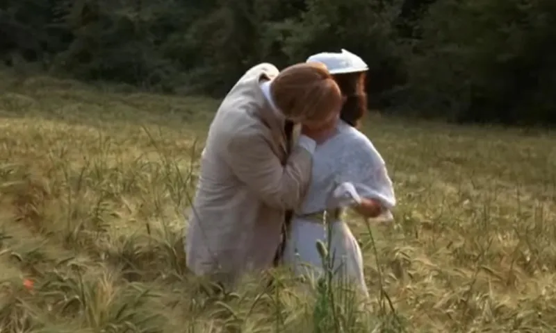A man and a woman in period costume, kissing in a poppyfield.